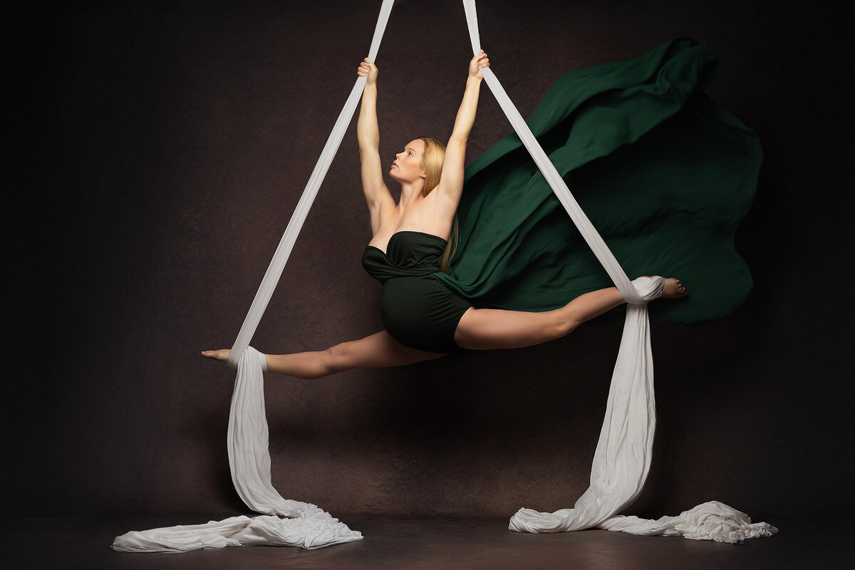 A graceful woman performs a stunning aerial split, suspended by white silk against a dark backdrop. The rich green gown flows elegantly as she holds her pose with powerful yet effortless precision. This captivating moment highlights the art of maternity photography in DFW.