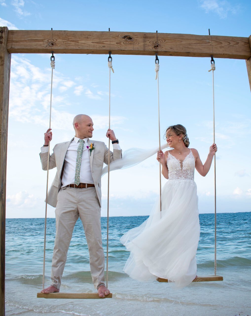 Bride and groom are standing on swings with the ocean in the background, looking at each other