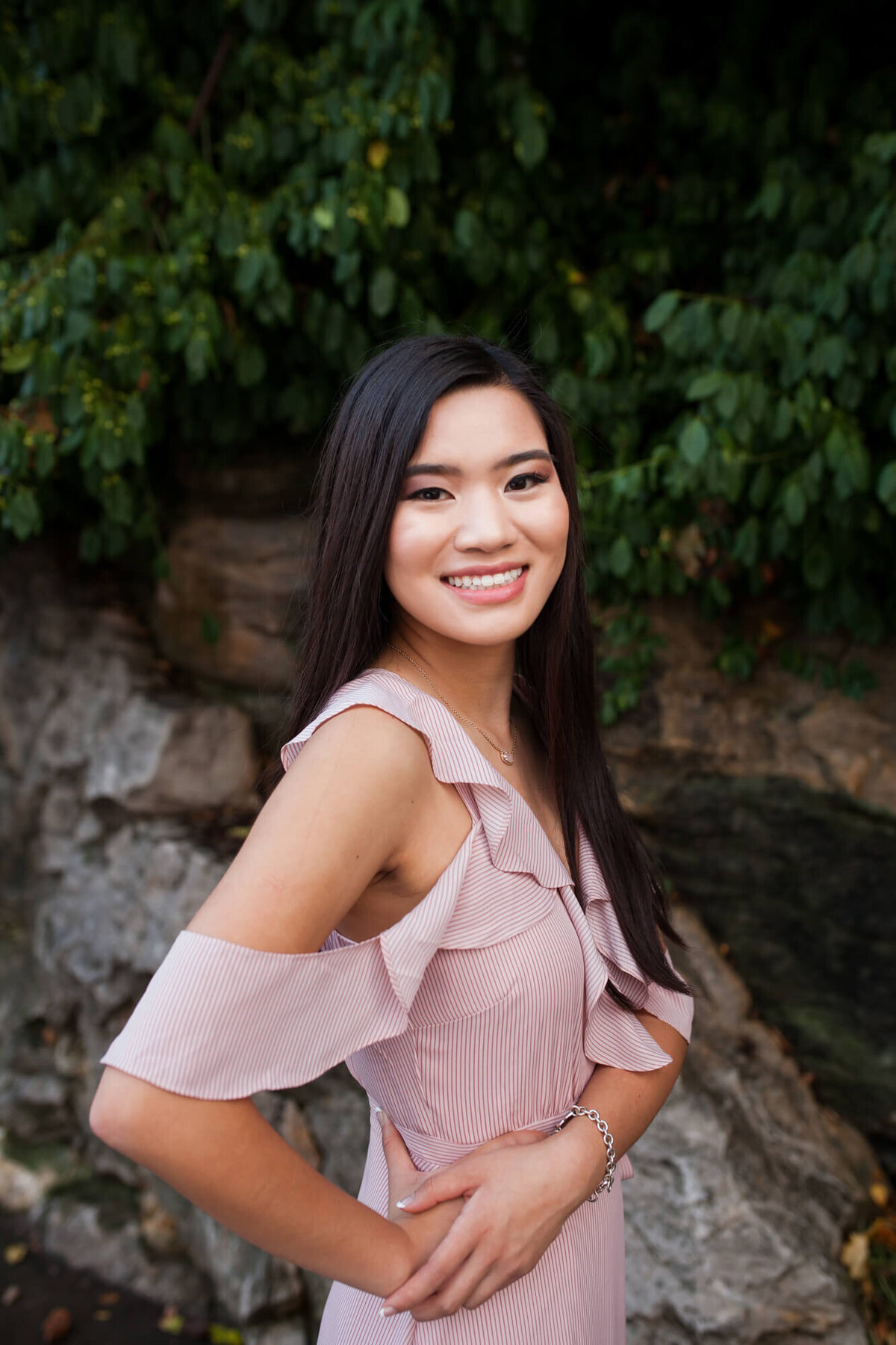 A lovely Asian American teen wearing a light pink cold shoulder dress poses smiling with her hands on one hip. Captured by Springfield, MO senior photographer Dynae Levingston.