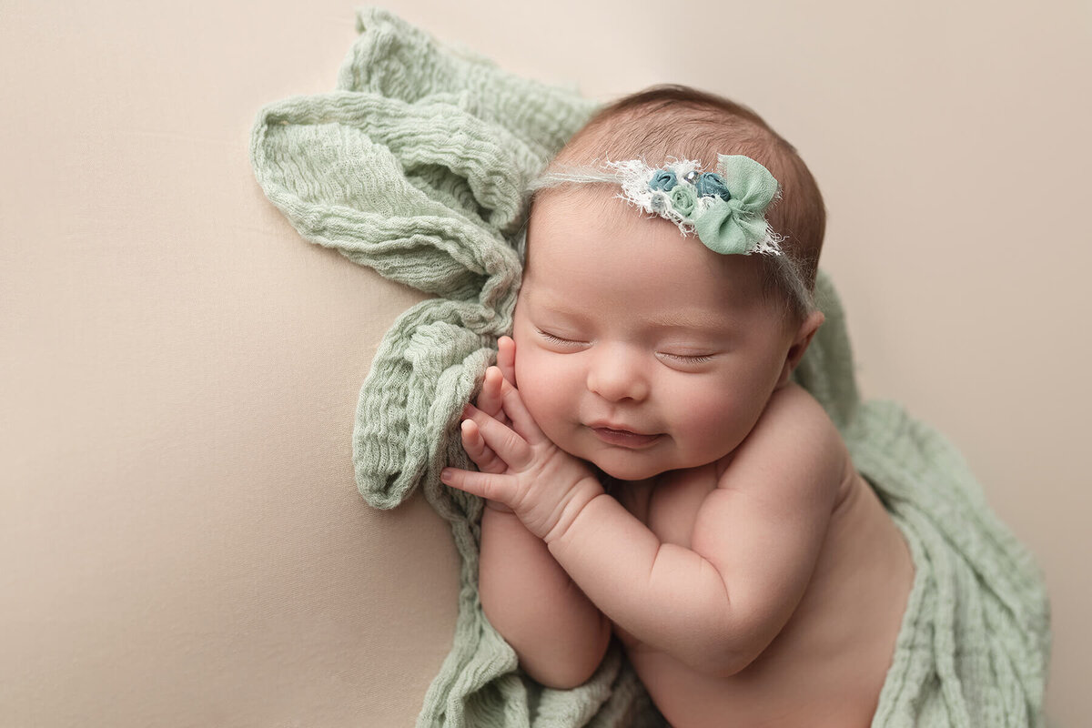 A newborn baby sleeps on her side while wrapped loosely in a green blanket with a matching bow