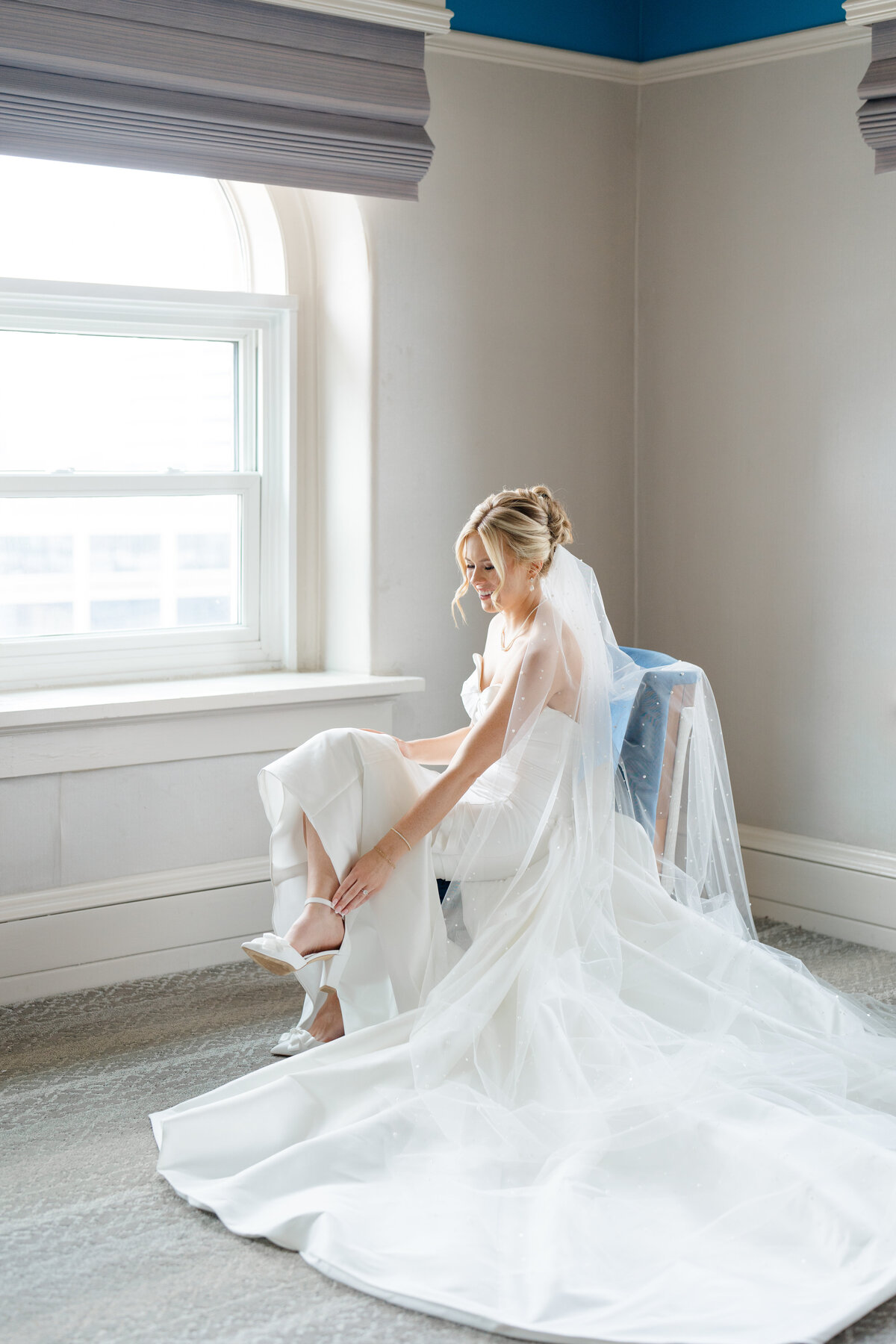 bride sitting on a blue chair in front of an open window putting on her bridal heels captured by Columbus wedding  photographer