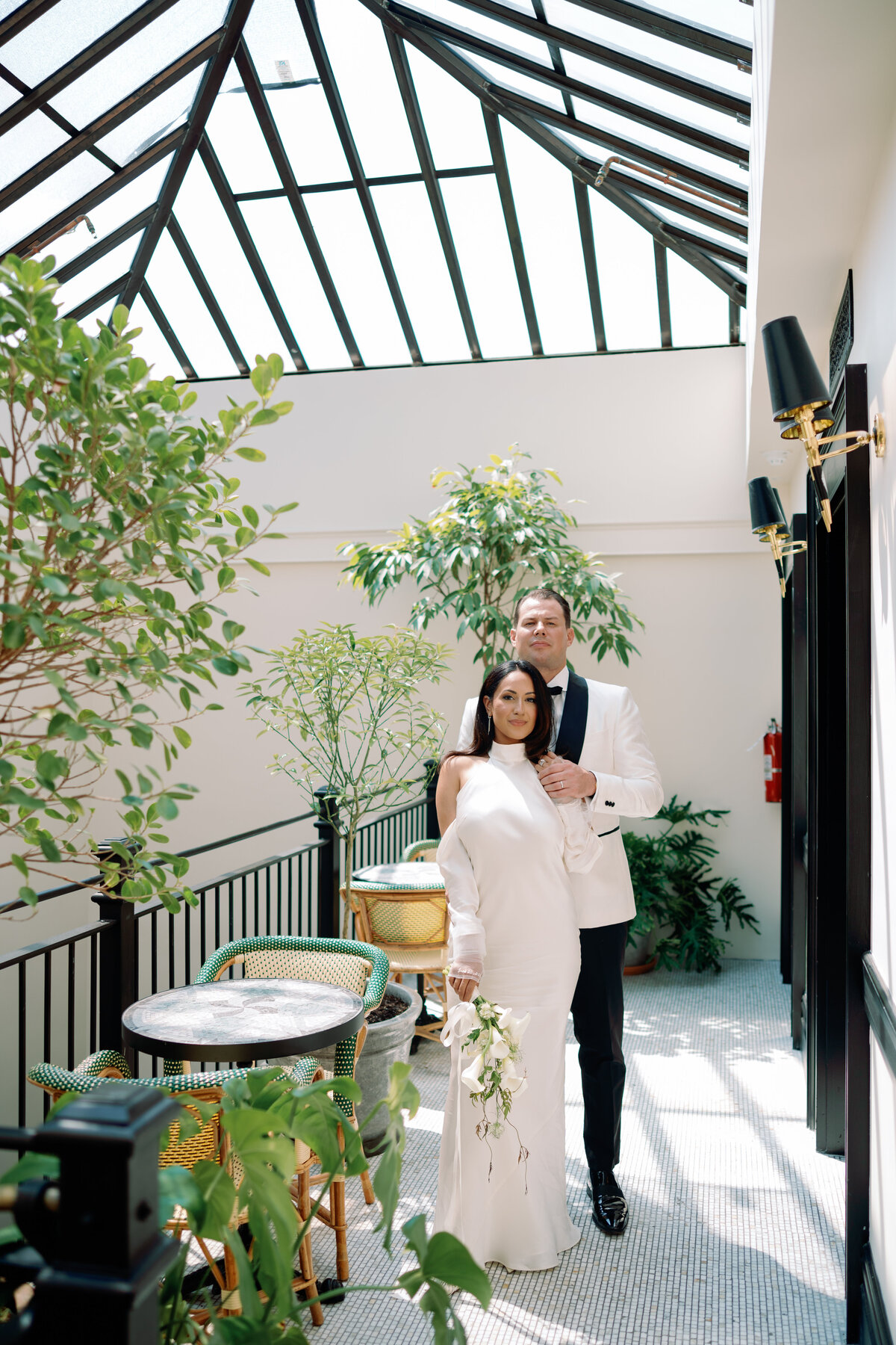 Couple walks hand in hand through a bright glass atrium surrounded by greenery on their wedding day.