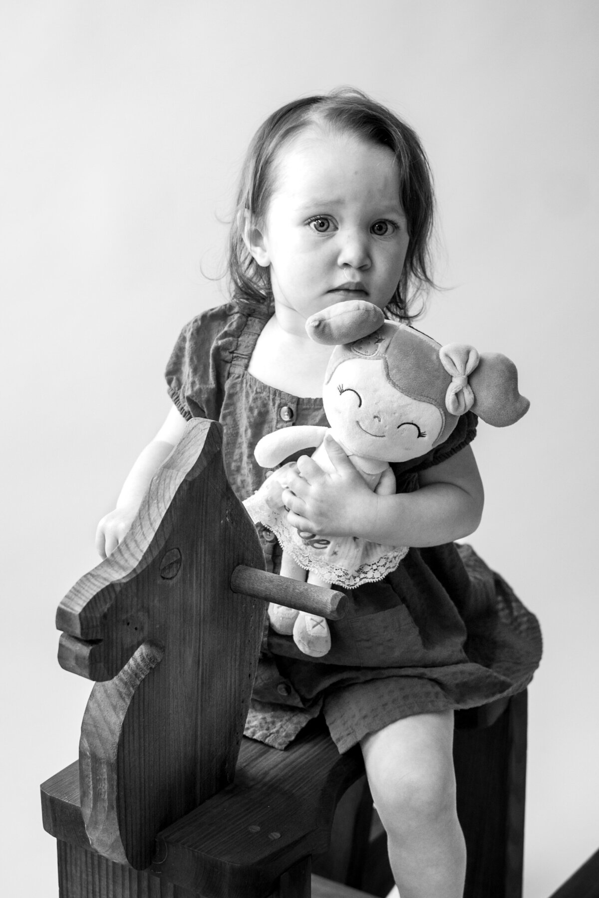 Black and white photo of girl sitting on wooden rocking horse holding a stuffed animal girl.