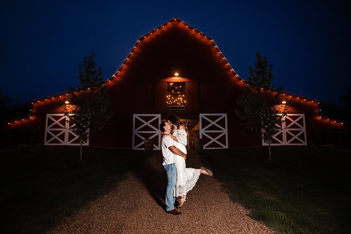 The groom lifts up the bride as he kisses her in front of a barn at night. The red barn has white detail around the closed windows and string lighting outlining the roof line. The groom is wearing an untucked white button down shirt and light blue jeans. The bride is wearing an off the shoulder lace boho maxi dress with a large lace ruffle around her shoulders. She has one foot kicked up in the air.