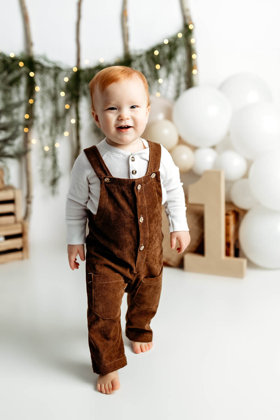 A smiling toddler with red hair, wearing brown overalls and a white shirt, walks barefoot on a white floor. Behind them, there are white and beige balloons, a large wooden number one, and a backdrop with fairy lights and greenery.