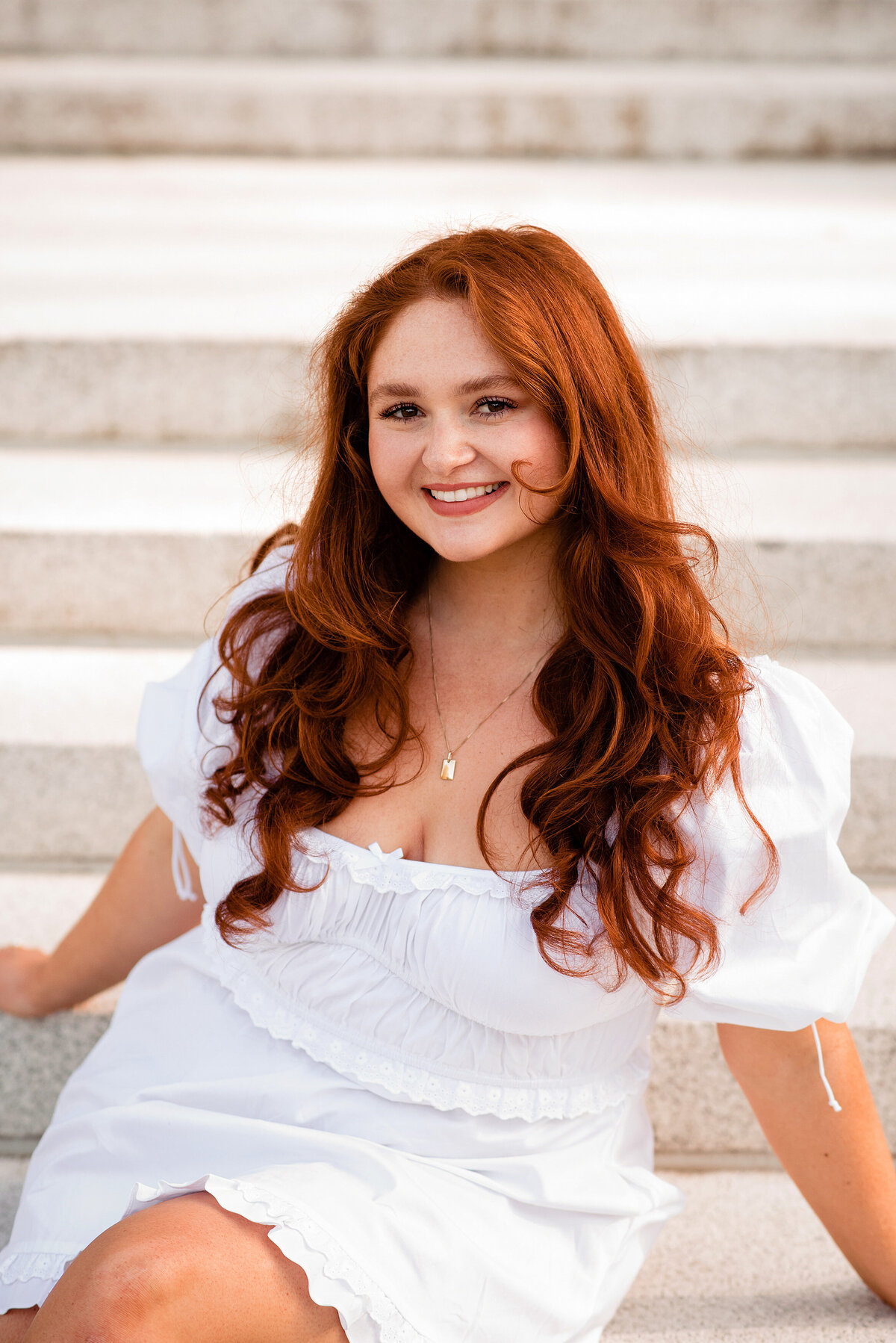 Senior Girl headshot smiling at the Camera wearing a white dress and sitting on the stairs