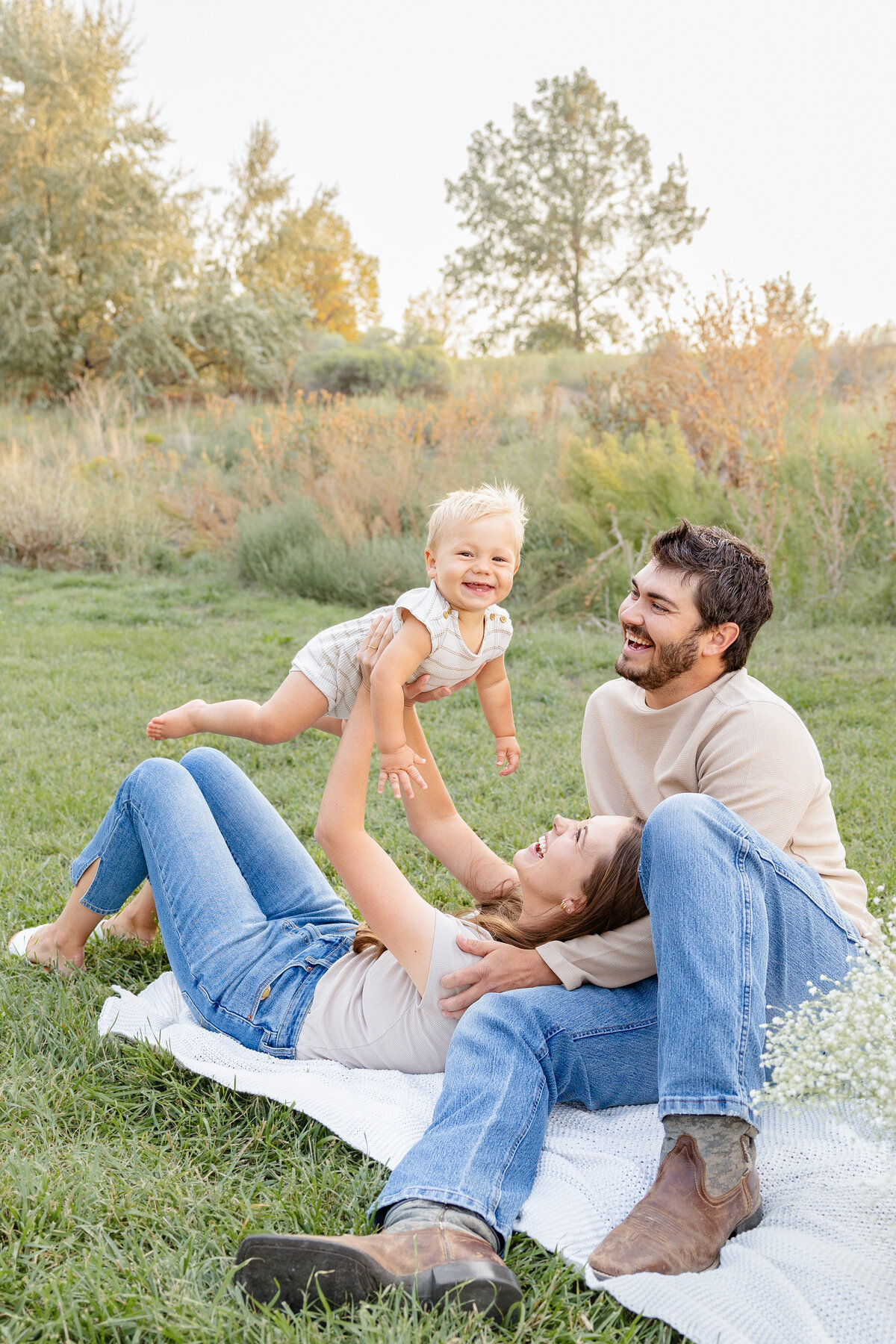 A baby giggles as his mother lifts him into the air, with both parents smiling brightly in a beautiful outdoor setting in Eagle Mountain, Utah.