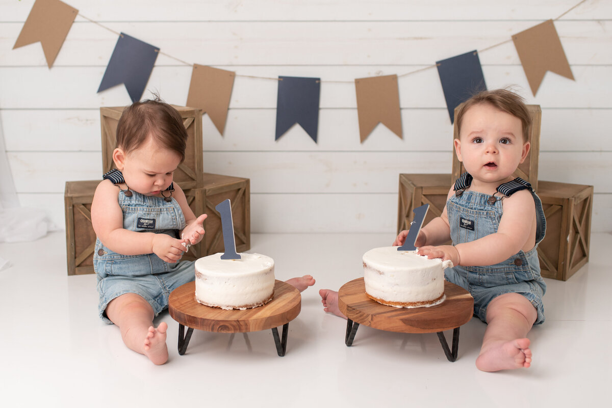 Twin boys looking at their cakes with blue and tan backdrop