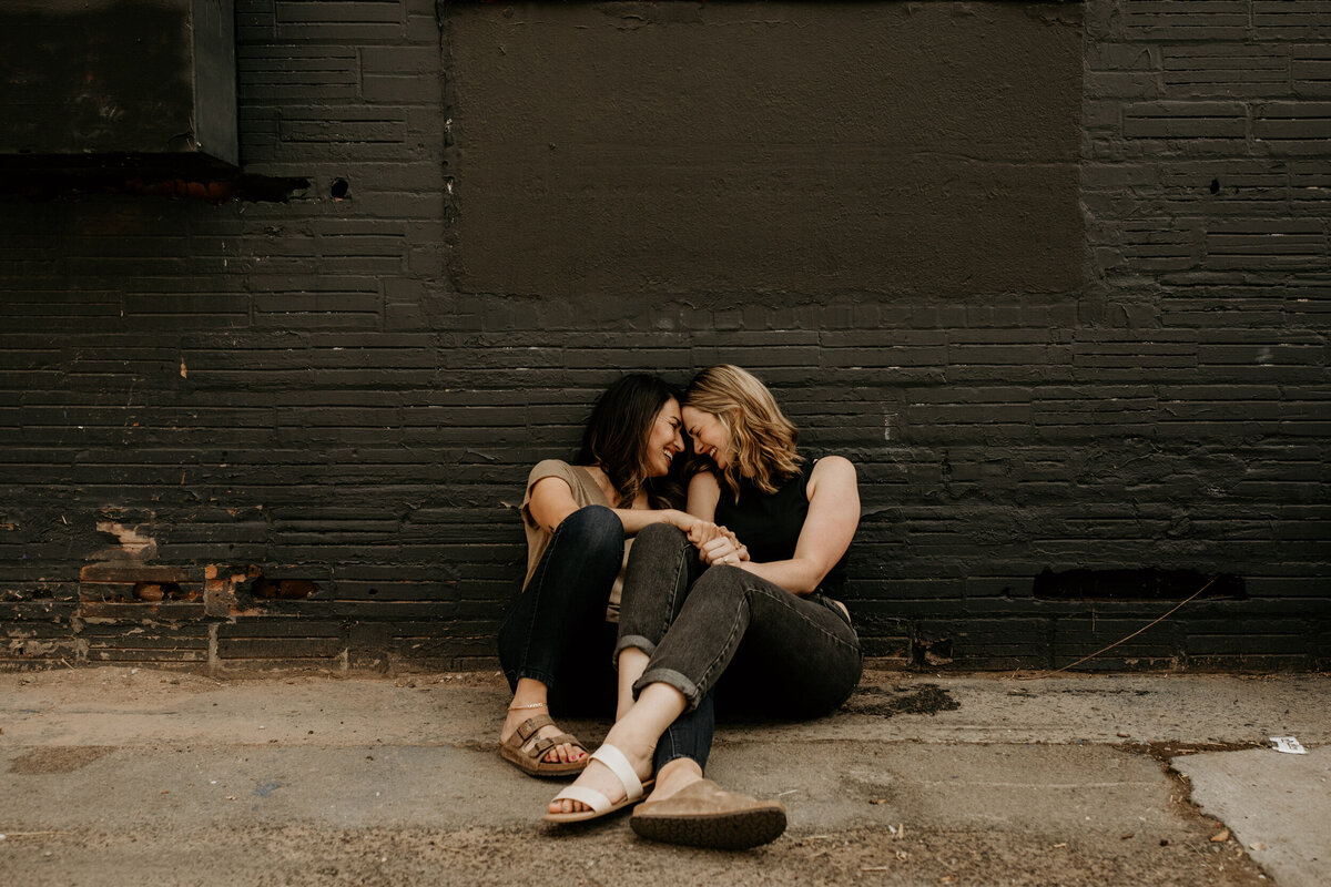lesbian couple sitting against a black wall in a alley in downtown Albuquerque