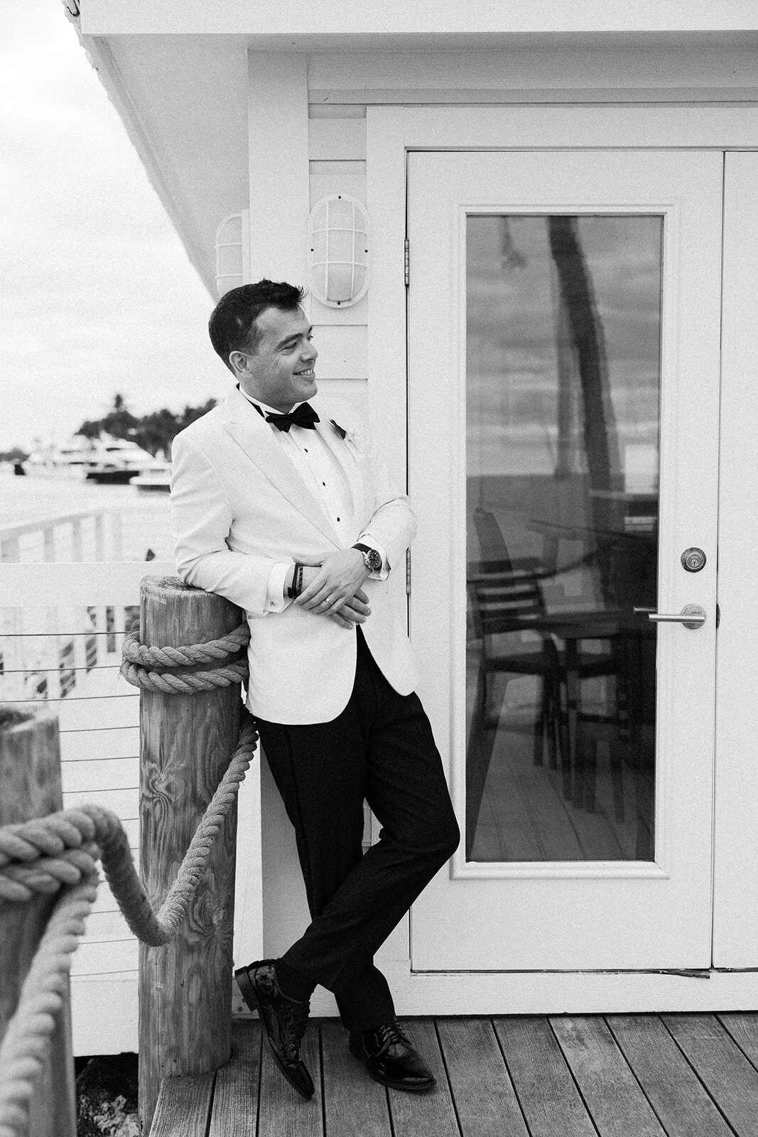 A classic black-and-white portrait of the groom leaning casually by the waterfront at Ocean Reef in Key Largo, captured by Claudia Amalia Photography.