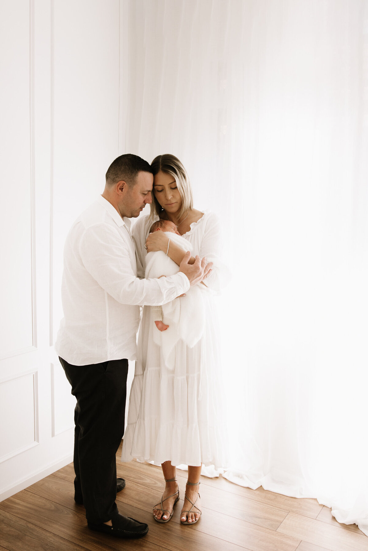 Full-length portrait of new parents gently holding their newborn baby in a softly lit, white studio in Southwest Sydney.