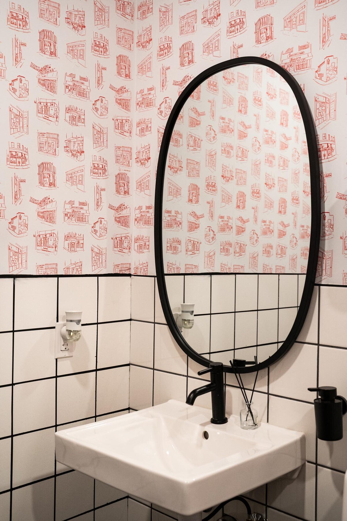 Small restroom with white square tiles, a black-framed oval mirror, and red illustrated wallpaper depicting hand-drawn storefronts.