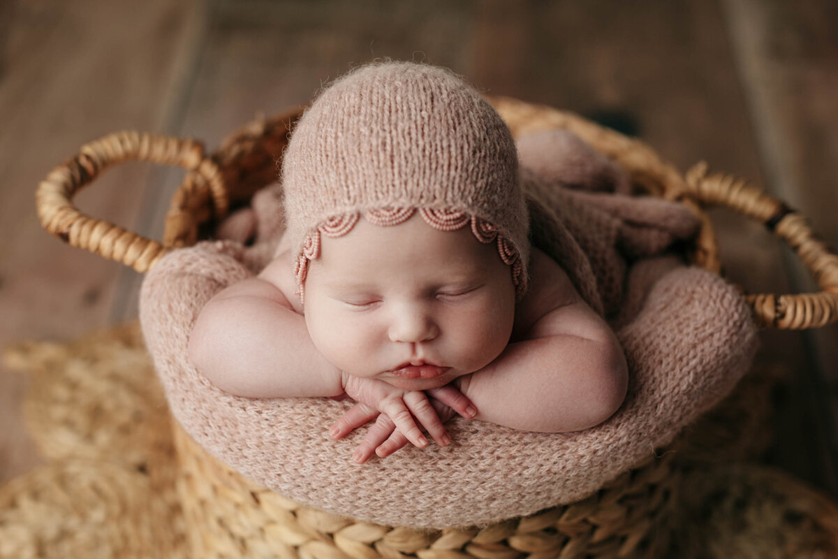 Studio newborn photography - sleeping baby girl in woven basket with pinky-taupe blanket flowing over the edge. Baby is resting her chin on folded hands and wearing a matching bonnet.
