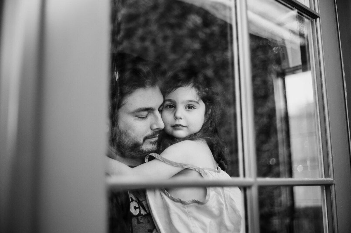 Black & white image shot through a window of a dad curled into his daughter's neck sharing a sweet moment.