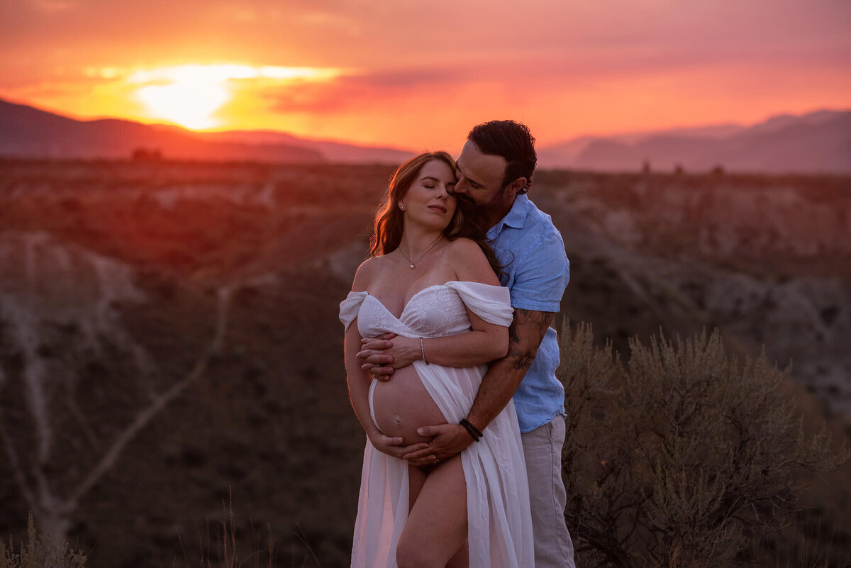 A couple embrace with a dramatic sunset in the background. Husband holds his wife from behind while she snuggles into him. Taken by Brittany Danielle Photography.