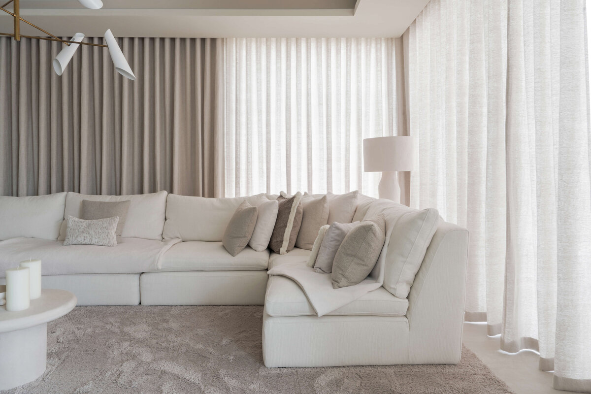 A spacious, modern living area. A large white couch with neutral throw pillows sits on a soft, textured cream rug. Behind the couch are floor-to-celiing cream drapes extending the length of the wall.