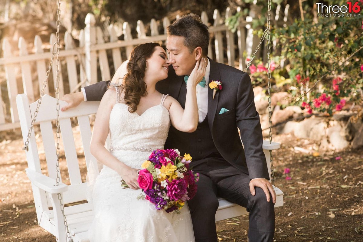 Newly married couple sit on a swing bench as the Bride leans in for a kiss