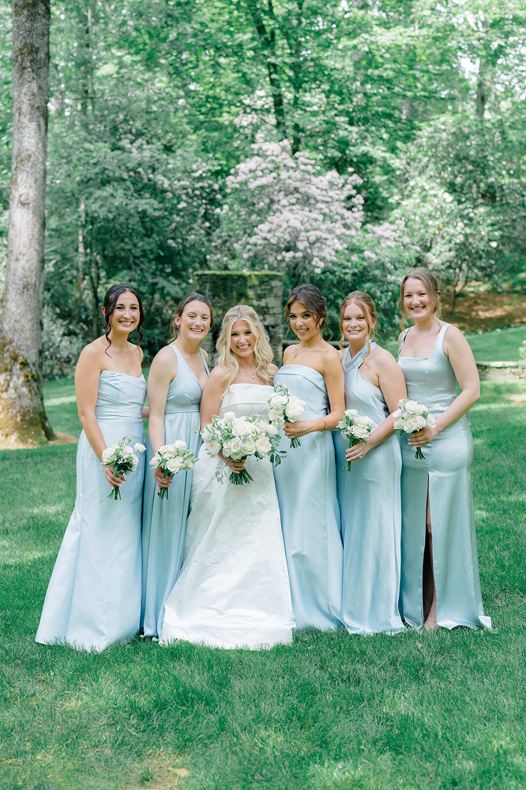 Bridal party standing together outdoors in Highlands, North Carolina, wearing light blue satin bridesmaid dresses and holding white floral bouquets.
