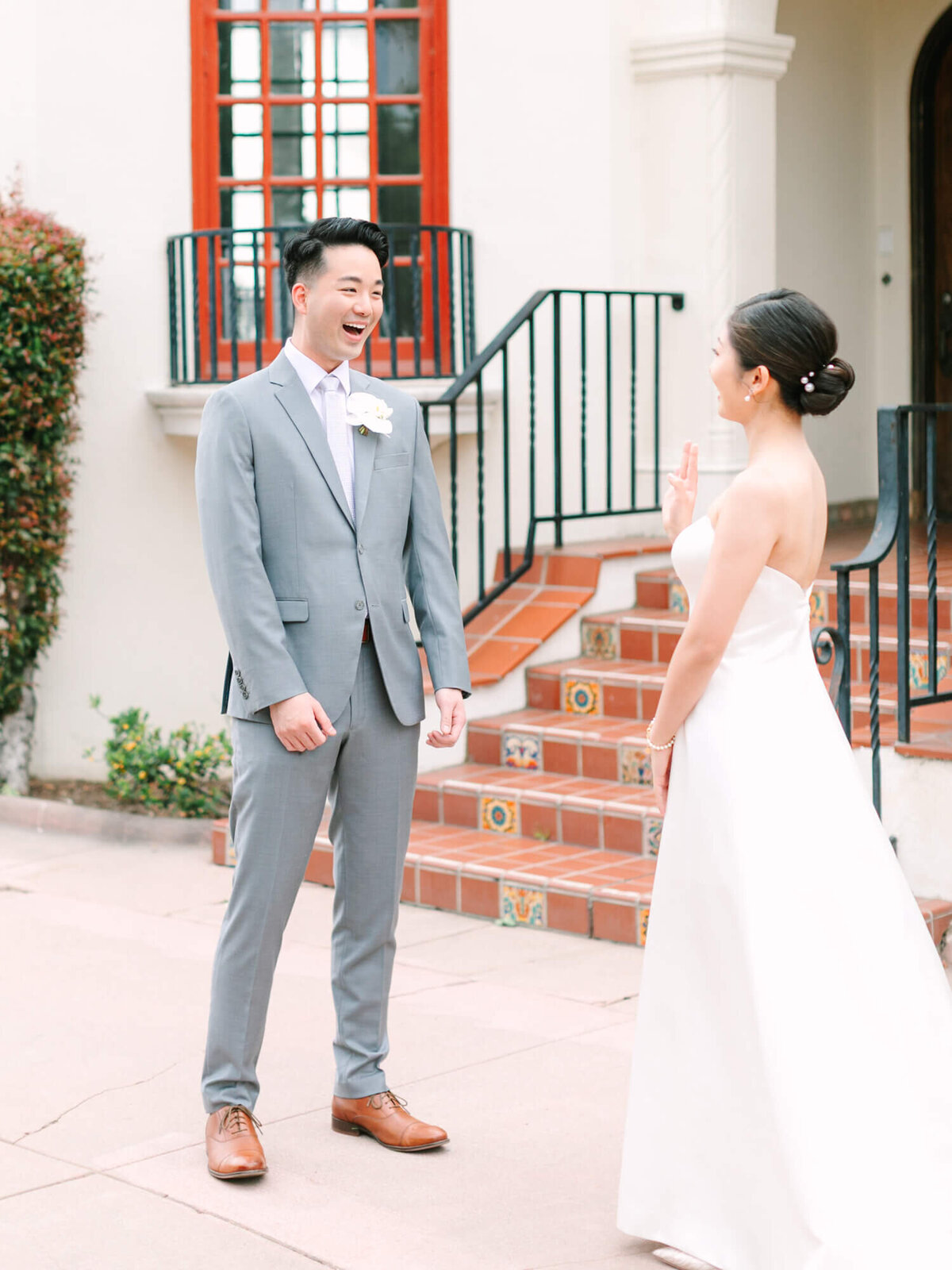 A bride and groom share a joyful moment outdoors. The groom in a gray suit looks surprised, while the bride in a white dress smiles. Elegant building in the background.