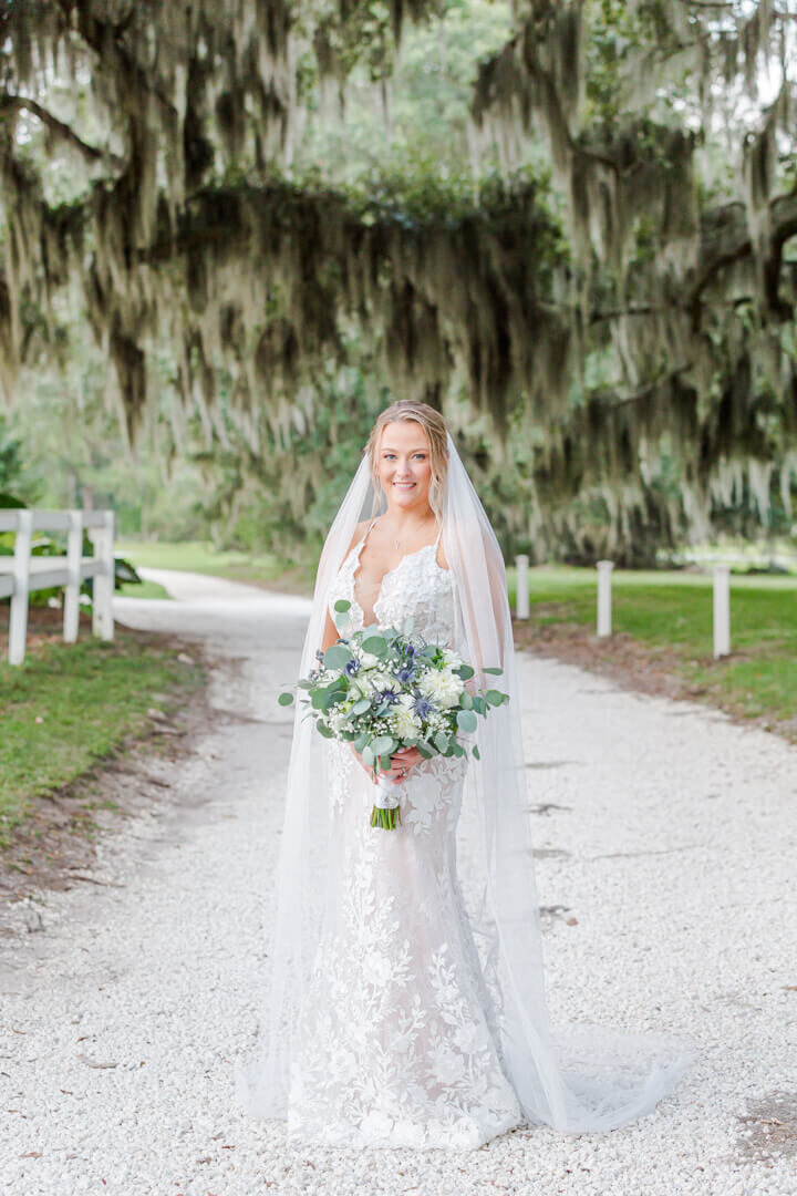 A smiling bride in a lace dress holds her bouquet under an oak canopy in a gravel path for an Arkansas wedding photographer