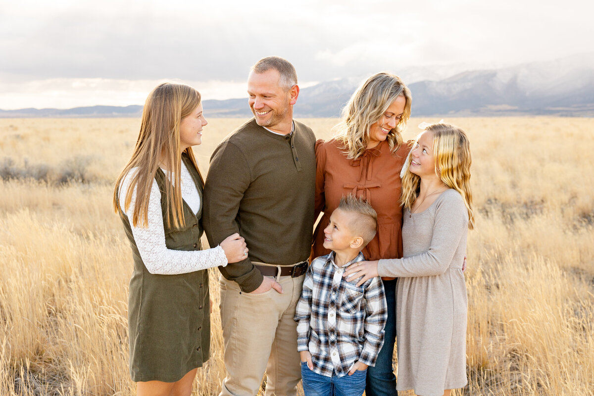 A family of five sharing smiles and warm glances as they embrace in a golden field. The parents look lovingly at each other while their children surround them with affection.