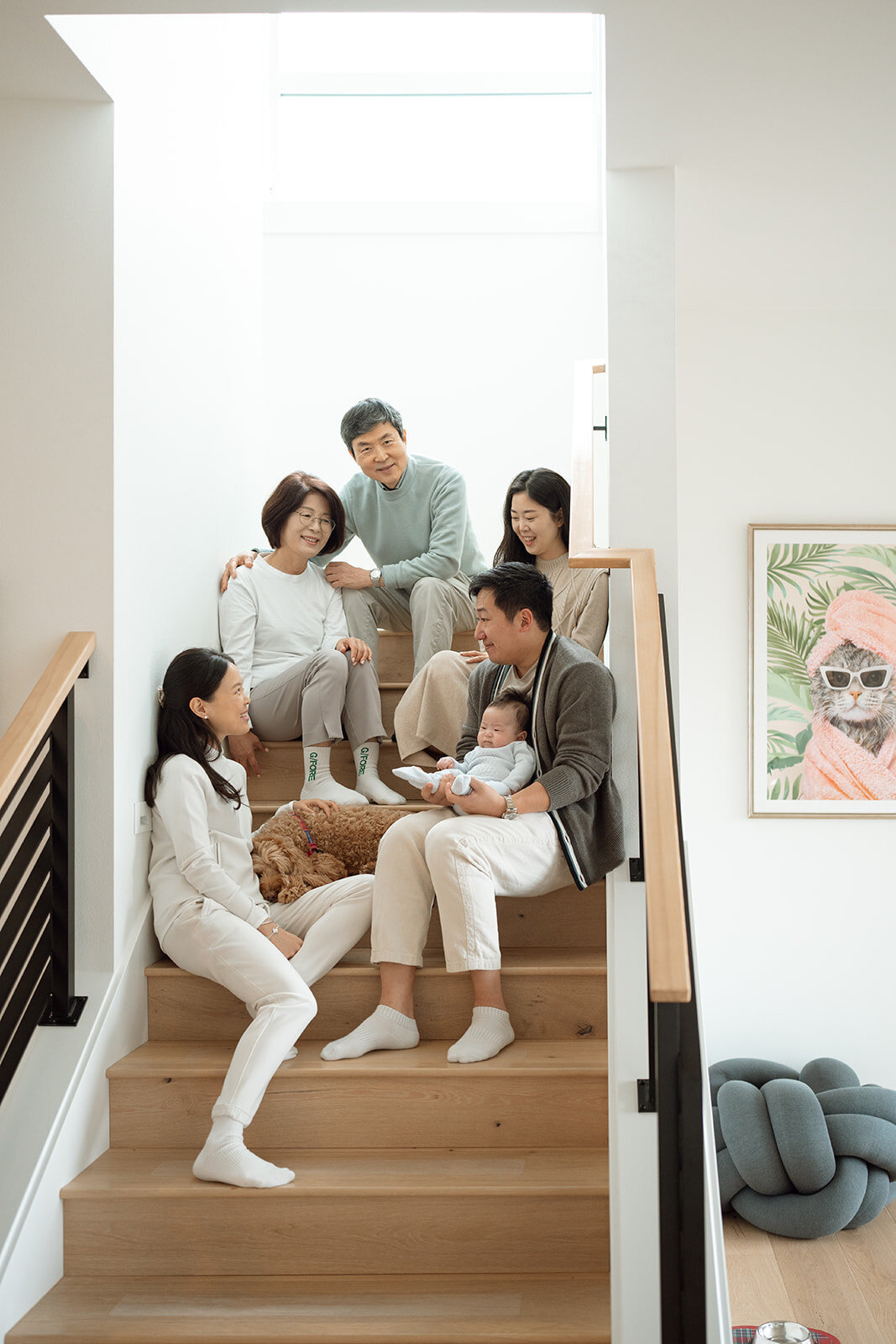 Multigenerational in-home family photography session in Irvine. Parents, grandparents, and a newborn sit together on a staircase with natural light, sharing relaxed and candid moments alongside their family dog.