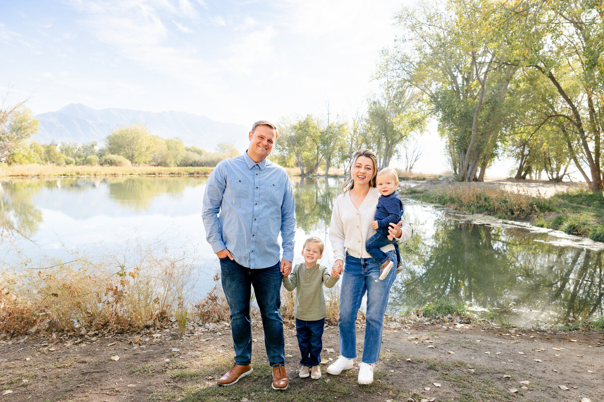 A young family stands near a reflective lake with mountains in the distance, holding hands and smiling, with the mother carrying their youngest child