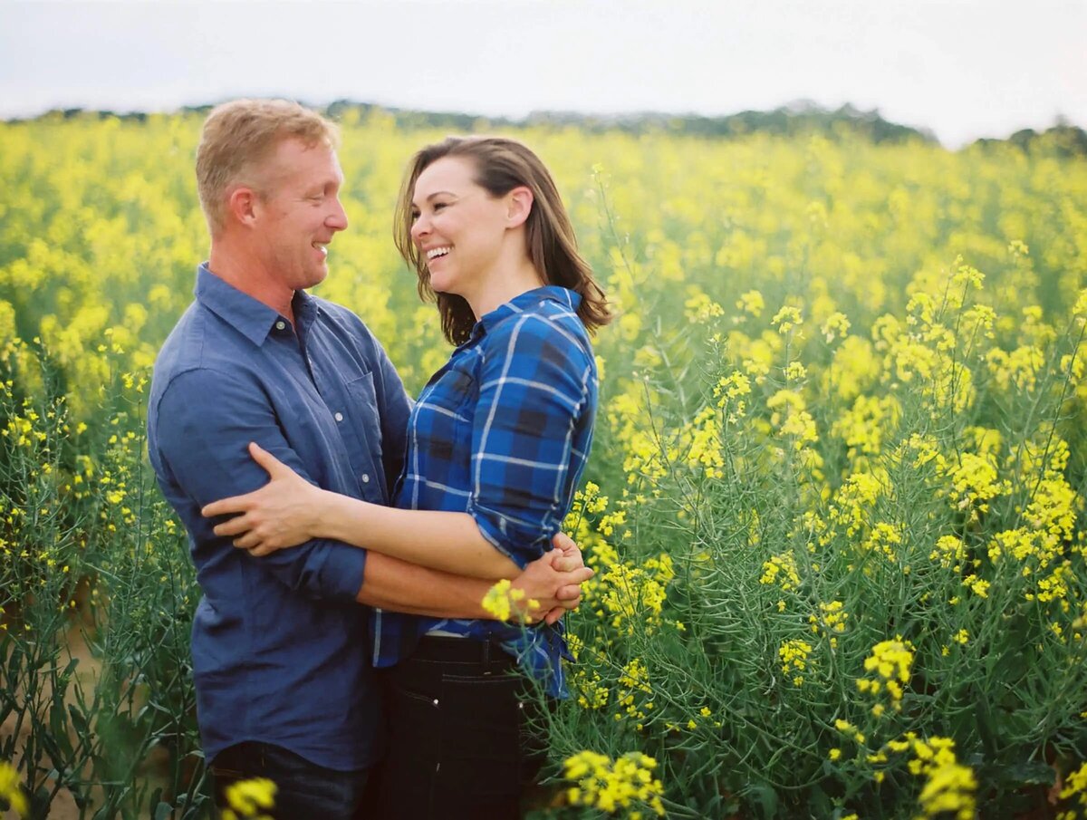 A couple stands in the midst of a vibrant yellow rapeseed field, sharing a loving embrace and a joyful moment.