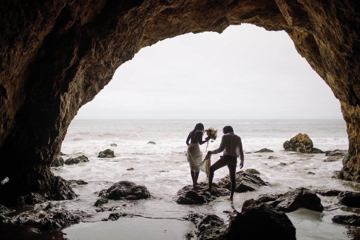 bride and groom step onto a rock in a sea cave