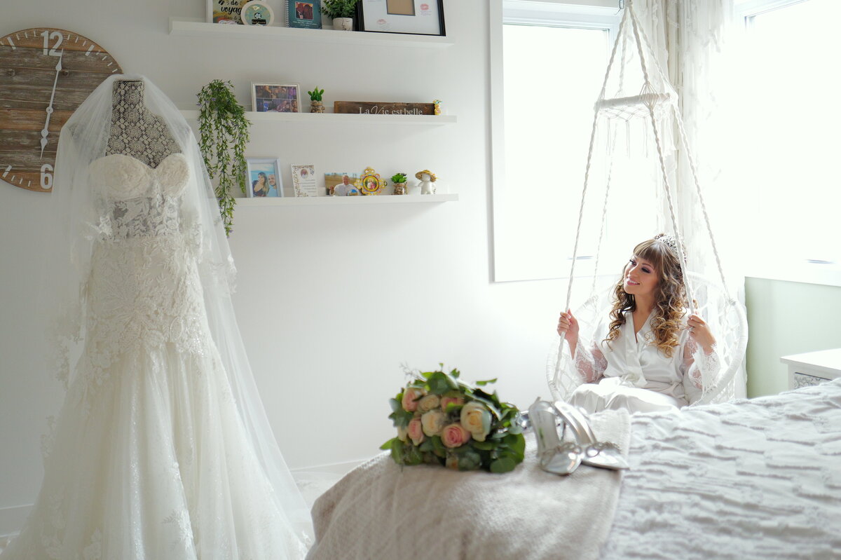 A bride sits gracefully in a swing, gazing at her wedding dress with admiration. This charming image captures a relaxed and whimsical moment, highlighting the bride’s elegance and the intricate details of her bridal gown in a unique and enchanting setting.