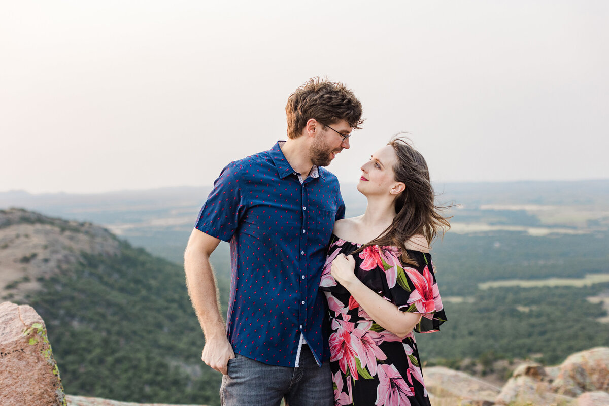 A couple holding each other close and looking into each other's eyes during their anniversary photoshoot in the Wichita Mountains in Oklahoma. The woman on the right is wearing a black dress covered in many colorful, large flowers while the man on the left is wearing a short sleeve blue dress shirt and dark jeans. A sprawling landscape can be seen behind them off into the distance.