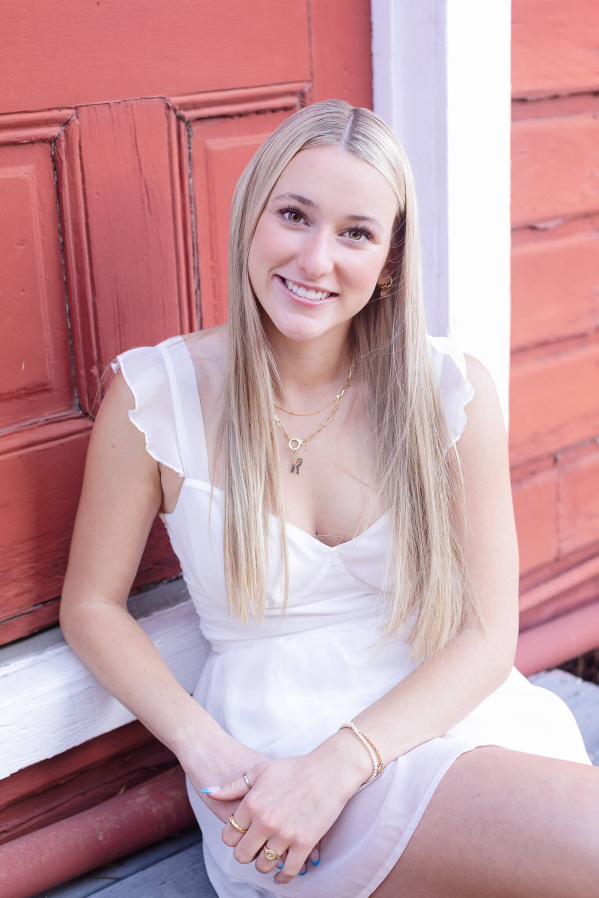 A young woman with long blonde hair sits smiling in front of a red wooden door. She is wearing a white dress and gold jewelry.