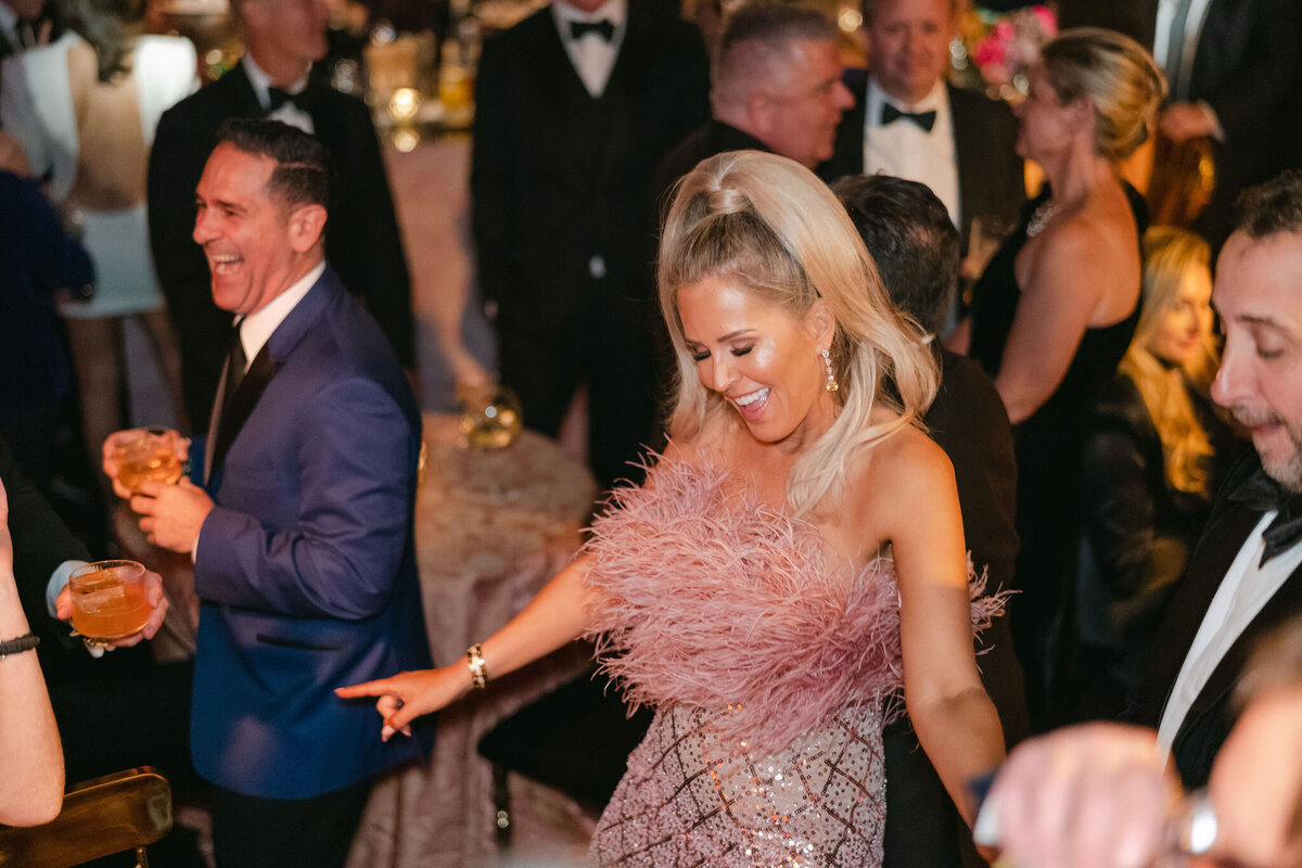This vibrant and festive photo was taken at a tented wedding in Virginia by the talented photographer Elisabetta Marzetti. In the center of the scene, a joyful woman in a feathered pink dress is captured celebrating and dancing exuberantly at this lively formal event. Surrounding her, elegantly dressed attendees are seen socializing, with some holding drinks in hand. The backdrop is adorned with sophisticated decor and warm lighting, enhancing the overall festive atmosphere of the gathering. Clayton Films was present as the videographers capturing the memorable moments of the event.
