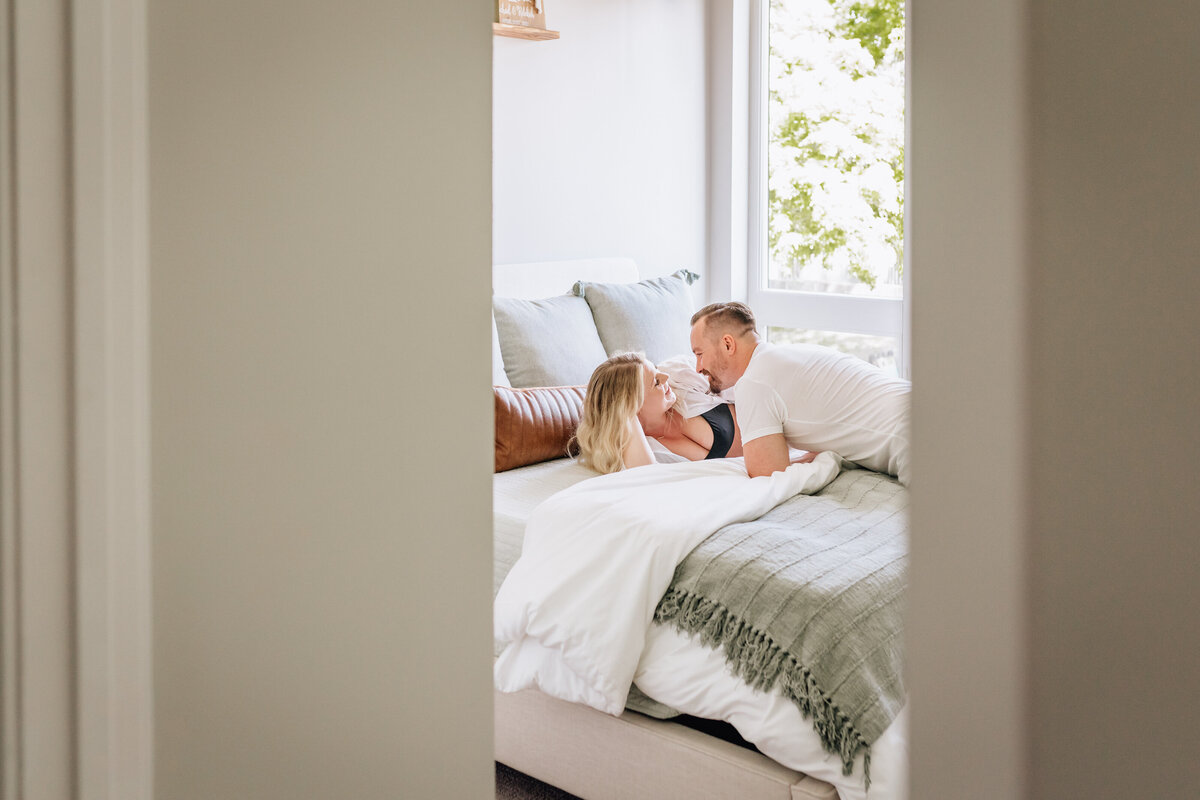 A glimpse through a doorway into a bedroom where the couple lies on a bed, smiling at each other in a tender moment, framed by soft natural light.