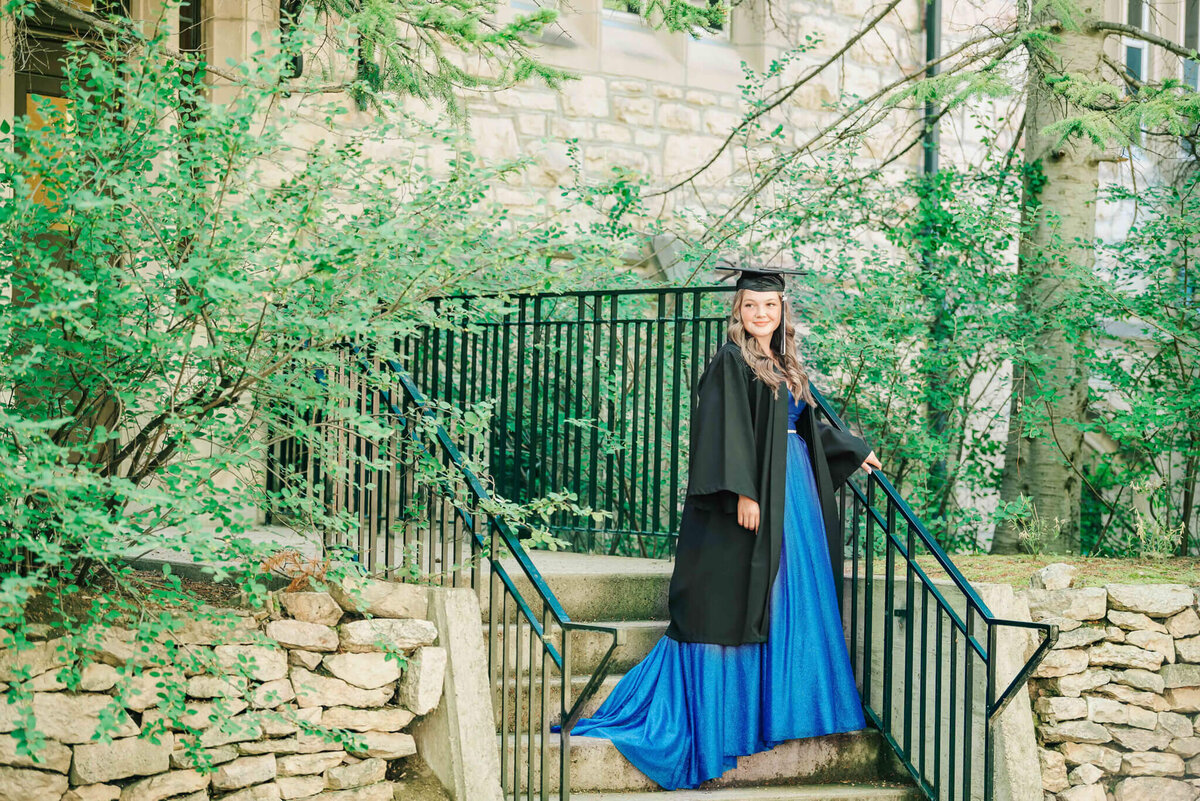 Girl in a blue dress, and grad gown, hat. Standing on stairs, with trees around. U of S campus.