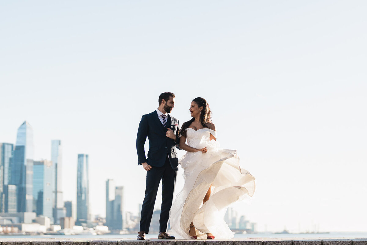Bride and groom standing on the Hoboken waterfront with the NYC skyline behind them as the bride’s dress blows dramatically in the wind during a joyful wedding portrait.