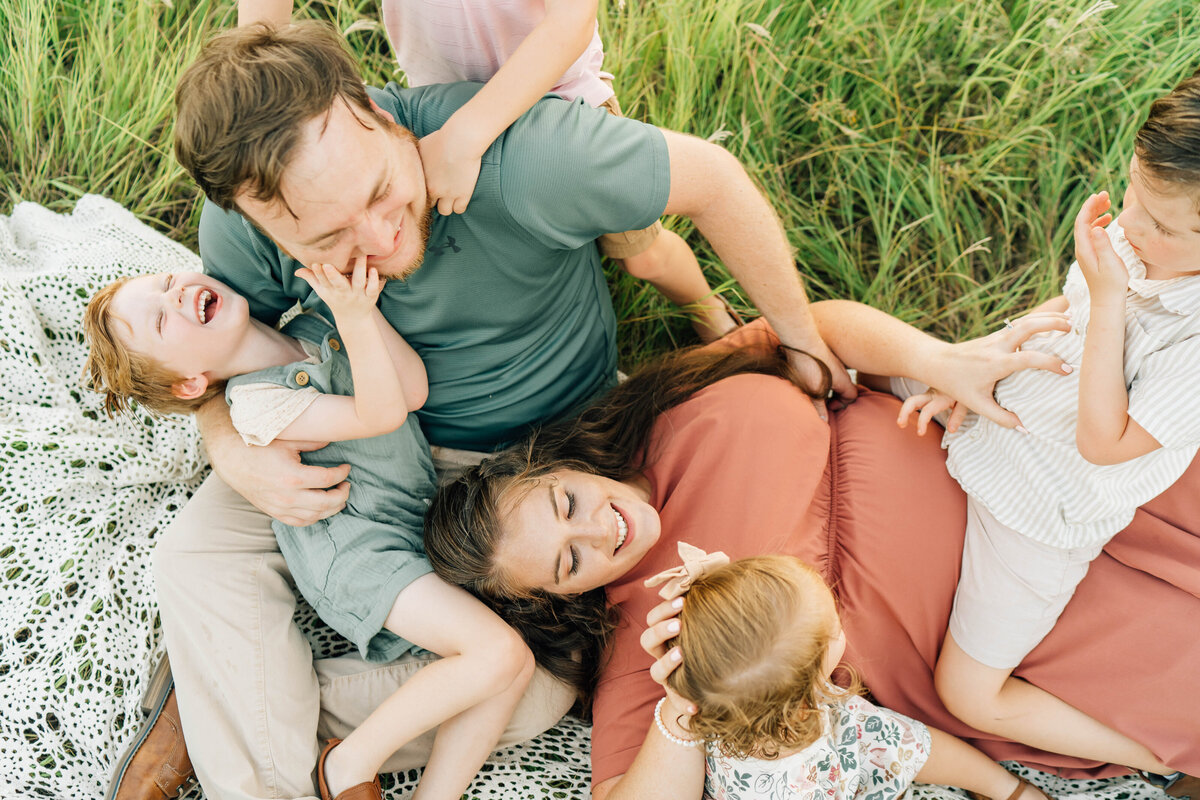parents laugh and tickle their four children while laying in a field