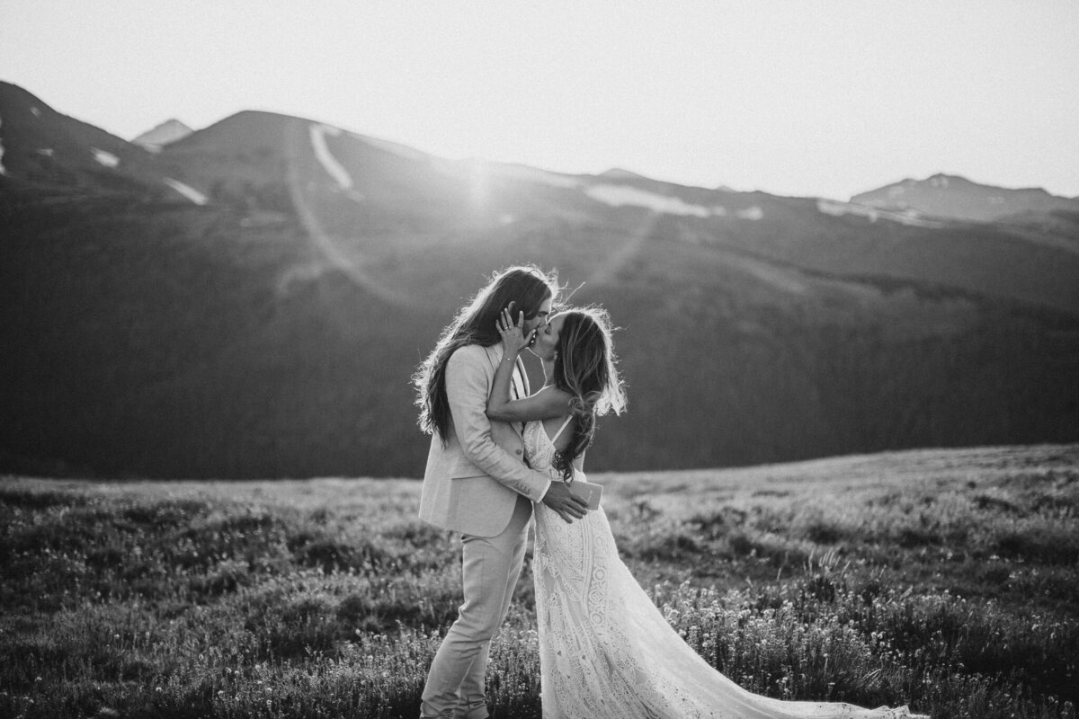 Black and white image of bride and groom wearing a suit and wedding gown kissing in a mountain field.