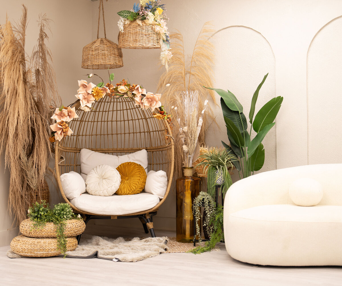 Cozy boho nook with a wicker egg chair adorned with flowers, soft cushions, pampas grass, and potted plants. Beige sofa adds warmth and relaxation.