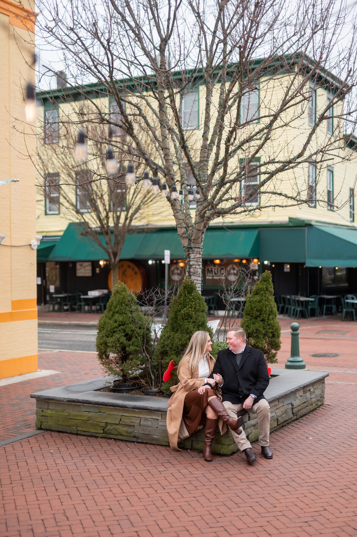 A man and woman sit together on a raised stone planter under a leafless tree in an urban outdoor area, with yellow buildings and green awnings in the background.