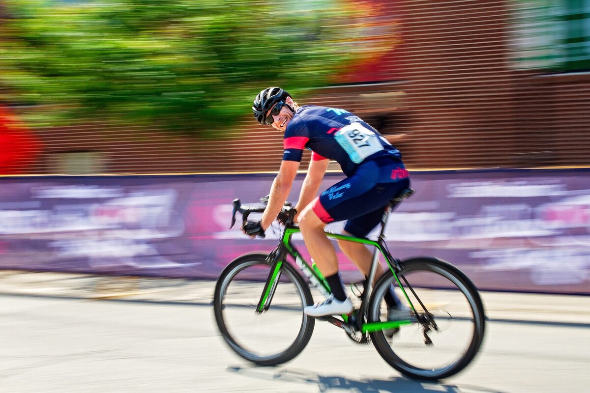 A cyclist on his bike races past while he looks at the camera. The background is blurry, indicating his high speed.