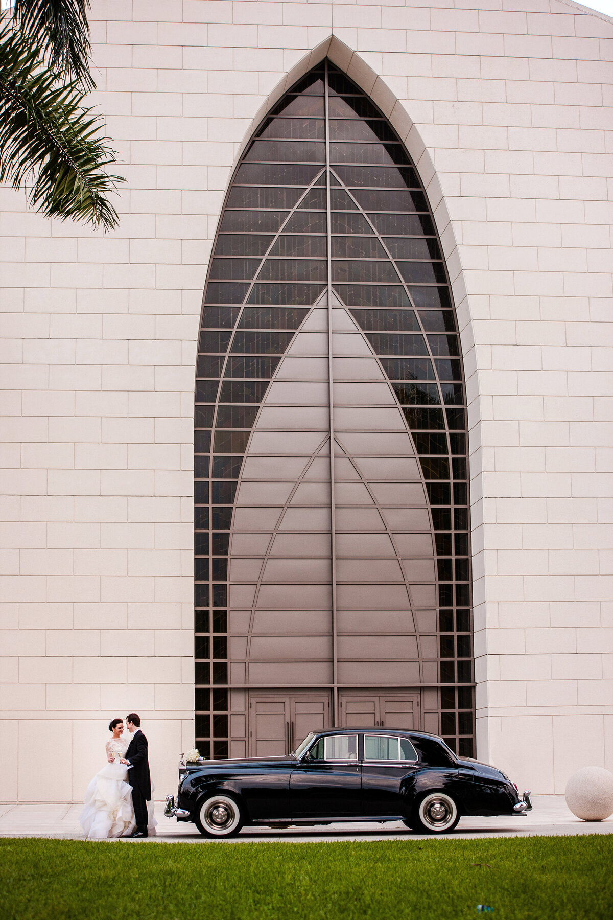 Bride and groom pose beside a classic car in front of modern Miami architecture.
