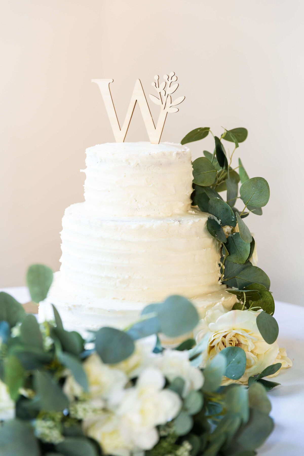 Three tiered wedding cake decorated with greenery with a W topper
