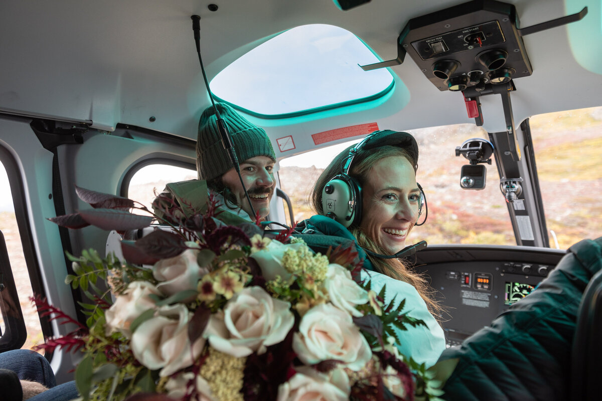 A bride and groom sit in the front of a helicopter smiling and looking back at the photographer while wearing headphones.