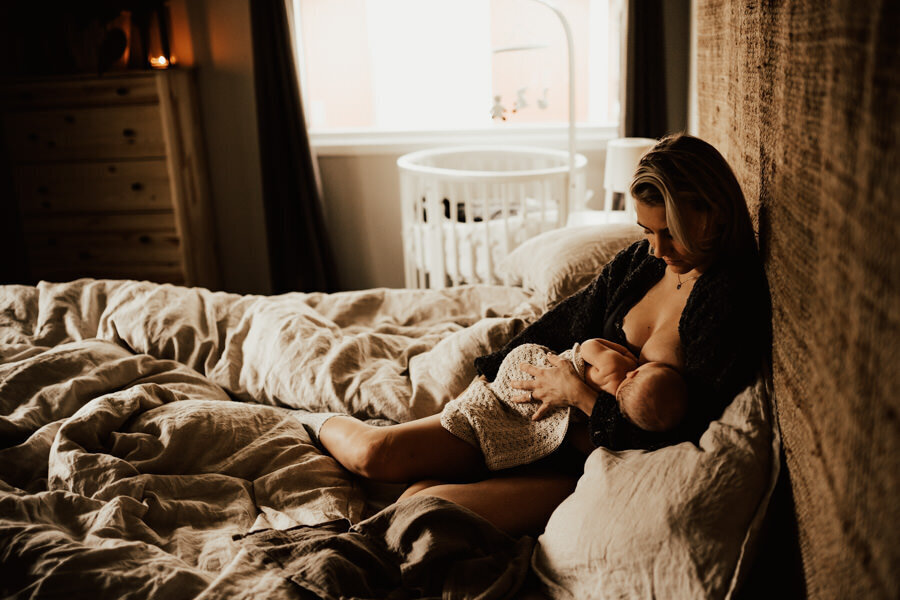 mamma sitter i bohemiskt sovrum med nyfödd bebis och ammar
