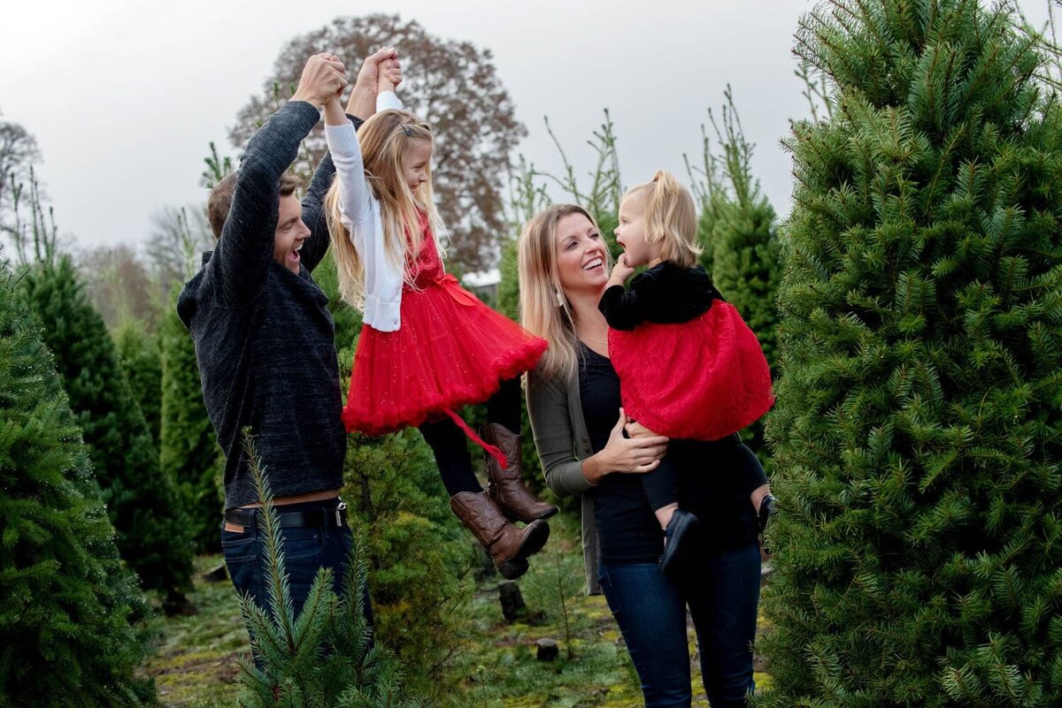 mom holding daughter in red christmas dress and dad holding other daughter by her arms while smiling at the tree farm.