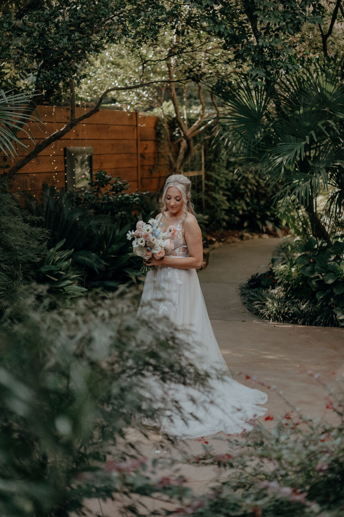 A bride holds her blue, white, and pink bouquet as she poses for a bridal photo surrounded by the lush greenery at the River Road and Jasmine House in Columbia, SC.