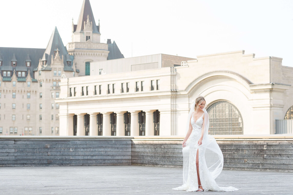 a brde with her dress billowing in the wind standing on the deck of the NAC with the Chateau Laurier wedding venue in the background.  Captured by Ottawa wedding photographer JEMMAN Photography