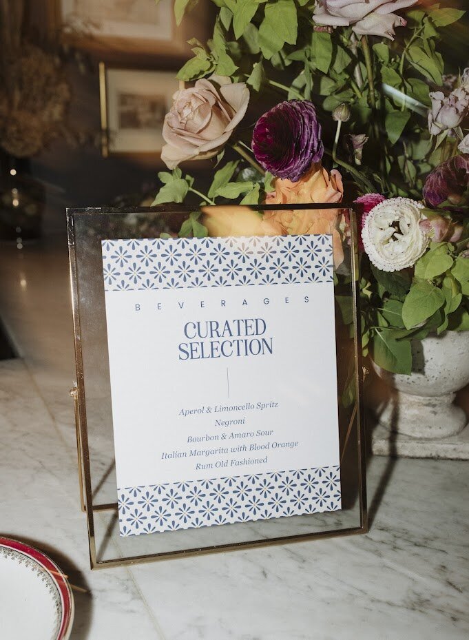 A framed "Curated Selection" cocktail menu in white with navy blue text and graphic pattern, highlighting the couple’s drink choices at their North & Navy wedding in Ottawa. The elegant menu sits in front of a floral arrangement on a marble bar counter, showcasing Frid Events' sophisticated wedding stationery design. Perfectly balancing style and personalization, this custom bar signage adds a modern yet timeless touch to the cocktail experience.