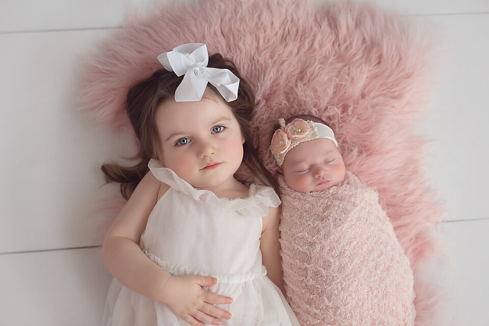 A toddler girl in a white dress lays alongside her sleeping newborn baby sister in a pink swaddle